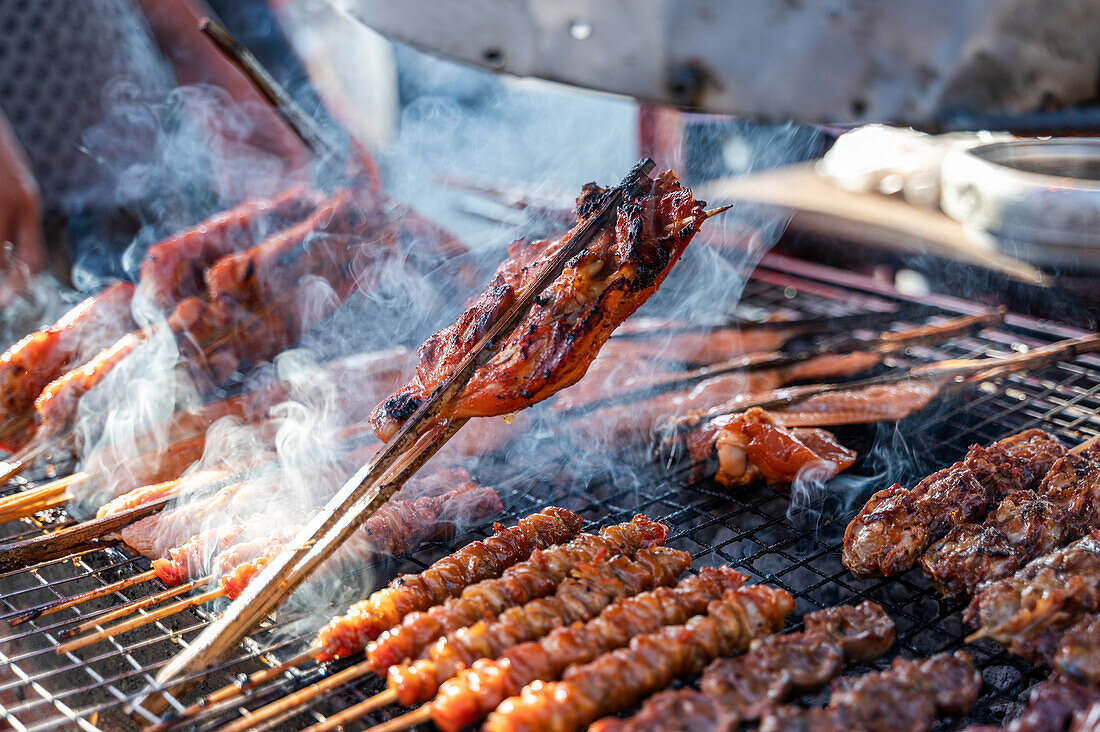 Juicy chicken skewers sizzle over charcoal, with smoke rising amid the busy food stall in Bangkok, Thailand, showcasing traditional street food.