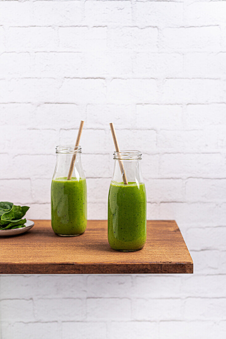 Two nutritious green smoothies in clear glass bottles with paper straws on a wooden shelf, against a white brick wall