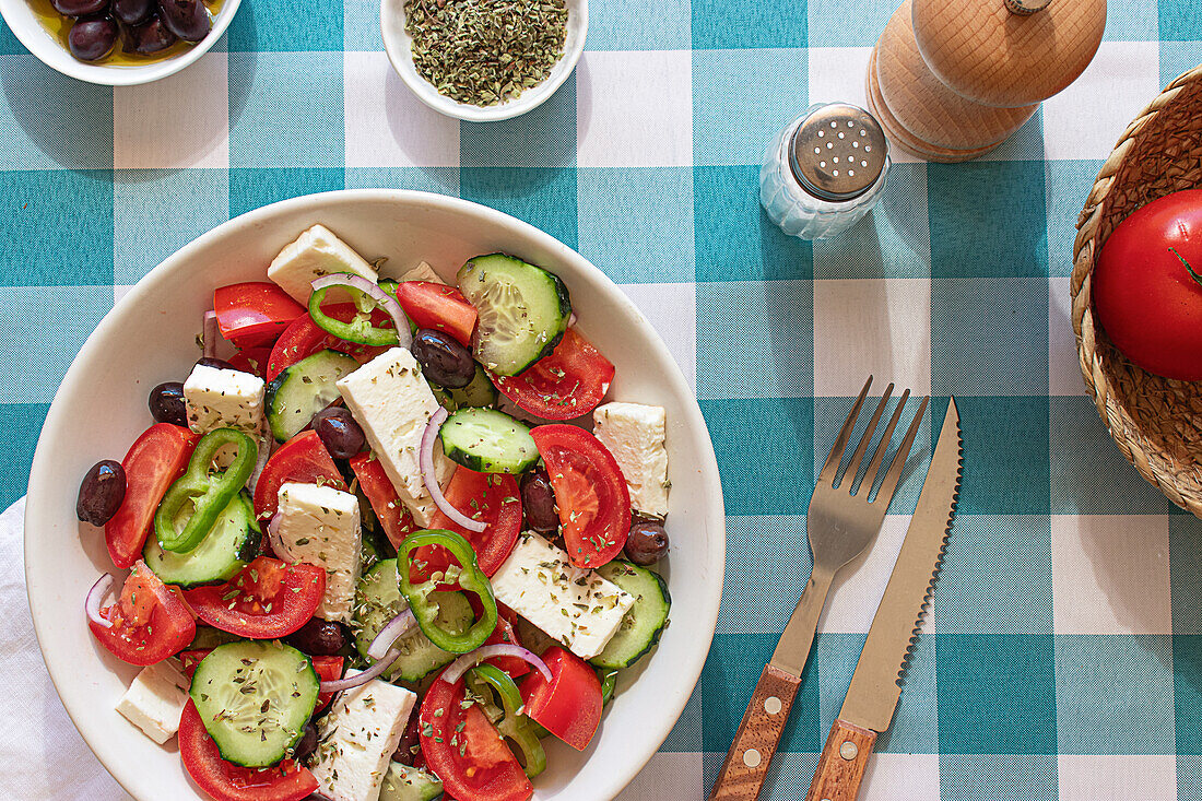 A colorful Greek salad with tomatoes, cucumbers, olives, feta cheese, and peppers, perfectly seasoned and arranged on a blue-and-white checkered tablecloth