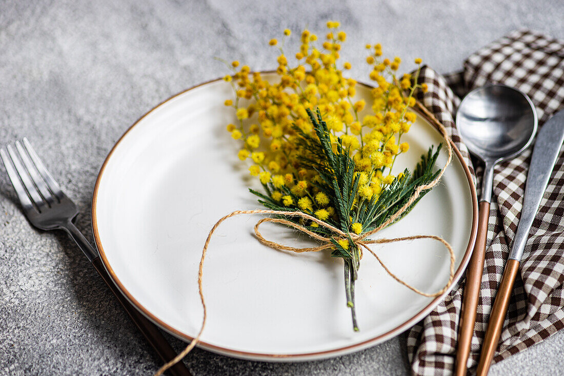 A rustic table setting featuring a white plate with a rim, accented with a sprig of mimosa flowers tied with twine, alongside silverware and checkered napkin