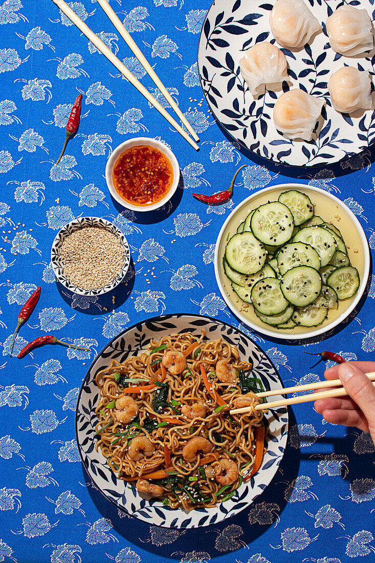 Top view of an assortment of Chinese dishes including shrimp noodles, cucumber salad, dumplings, and sauces. A hand is seen picking up noodles with chopsticks, enhancing the lively dining atmosphere