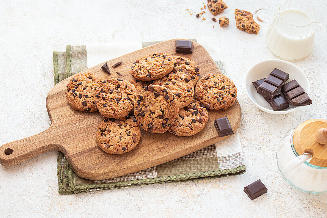 A tempting chocolate chip cookies on a wooden board, with chocolate chunks, sugar bowl, milk jar in the background Ideal for dessert and snack moments