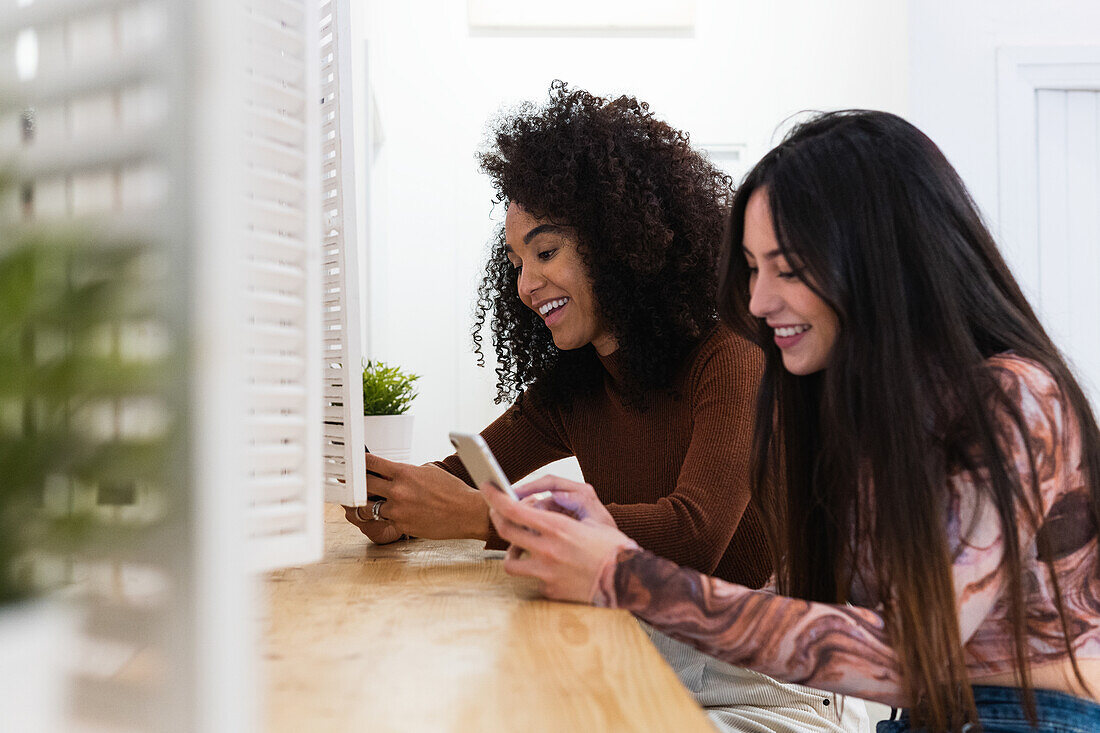 Seitenansicht von verschiedenen Freundinnen, die am Tresen einer Bar sitzen und Smartphones benutzen, während sie das Wochenende zusammen genießen