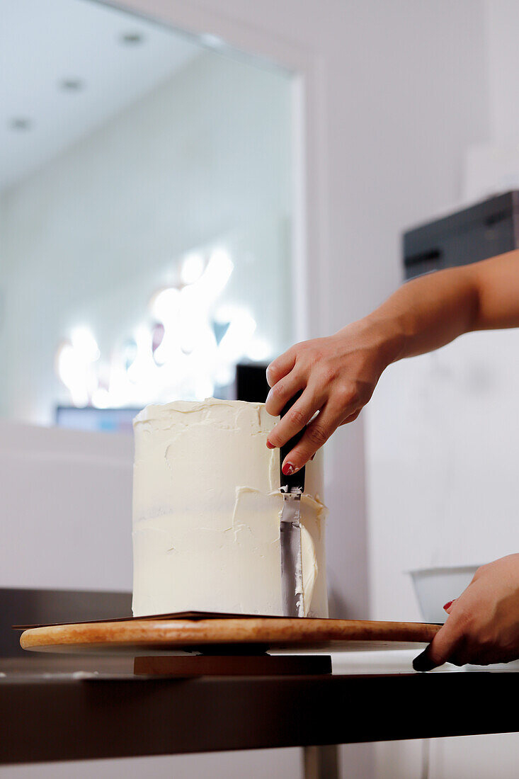 Eine Person trägt fachmännisch glatten Zuckerguss auf einen hohen Kuchen in einer modernen Kücheneinrichtung auf und demonstriert dabei Techniken zur Kuchendekoration