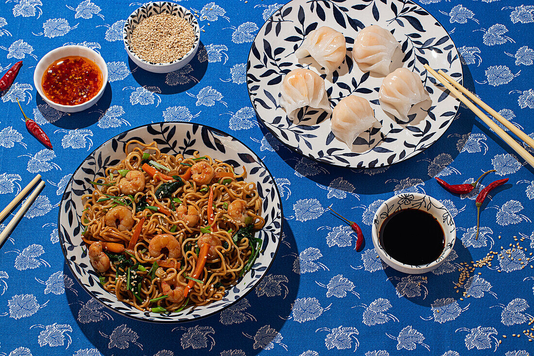 From above, a vibrant display of Chinese food on a blue floral tabletop with shrimp noodles, steamed dumplings in a patterned bowl, and condiments like soy sauce and chili oil