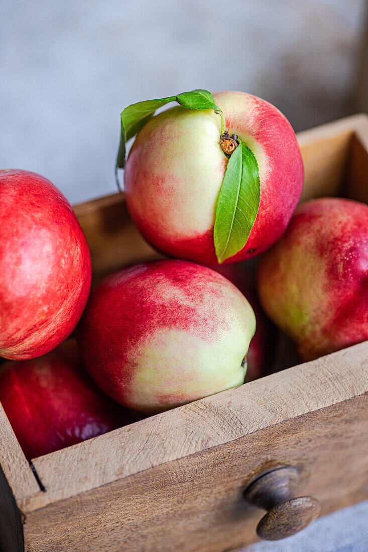 A close-up image showcasing fresh, ripe nectarines with vibrant red hues, nestled in a quaint wooden crate. Each nectarine features a tender green leaf, adding a touch of natural beauty.