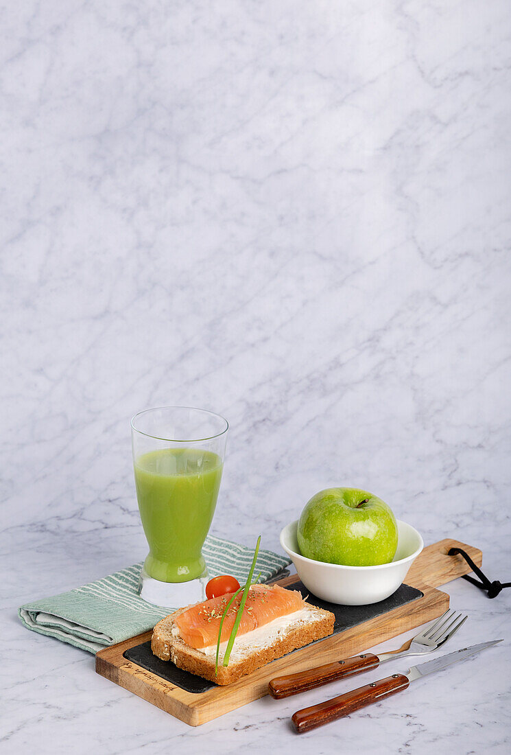 Healthy and appealing meal setup including a glass of cucumber and apple juice, green apple, and a toast topped with salmon and cherry tomato on a marble background.