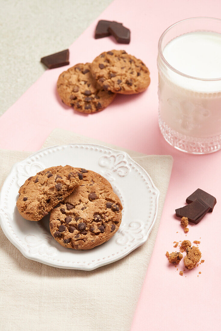 Top view of delicious chocolate chip cookies on a white plate with a glass of milk and scattered chocolate pieces on a pink background