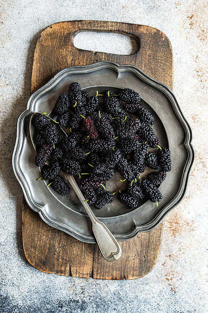 Top view of a rustic setting showcases fresh juicy mulberries in a vintage silver tray, resting on a weathered wooden chopping board, complemented by a textured silver spoon and a dusty backdrop.