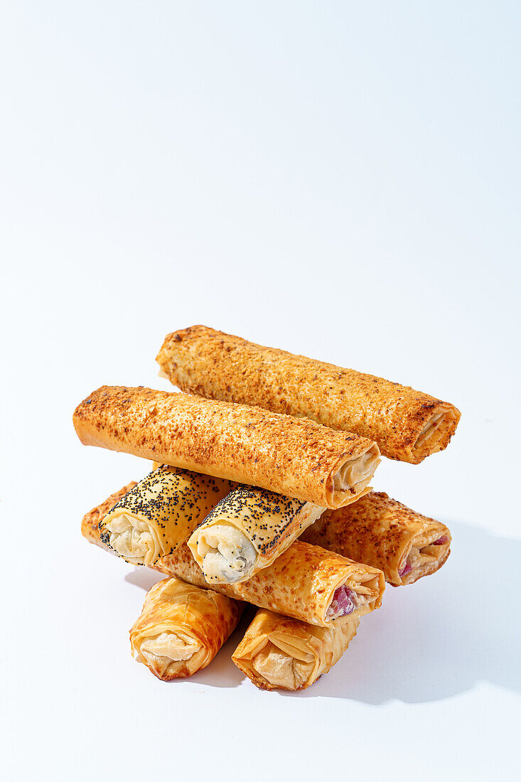 A variety of freshly baked puff pastry rolls, some topped with sesame and poppy seeds, arranged on a clean, white background.