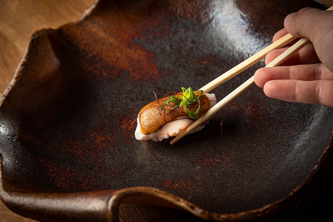 Nahaufnahme einer Hand, die ein sorgfältig zubereitetes Sushi-Stück mit Stäbchen auf einem dunklen, rustikalen Keramikteller zupft