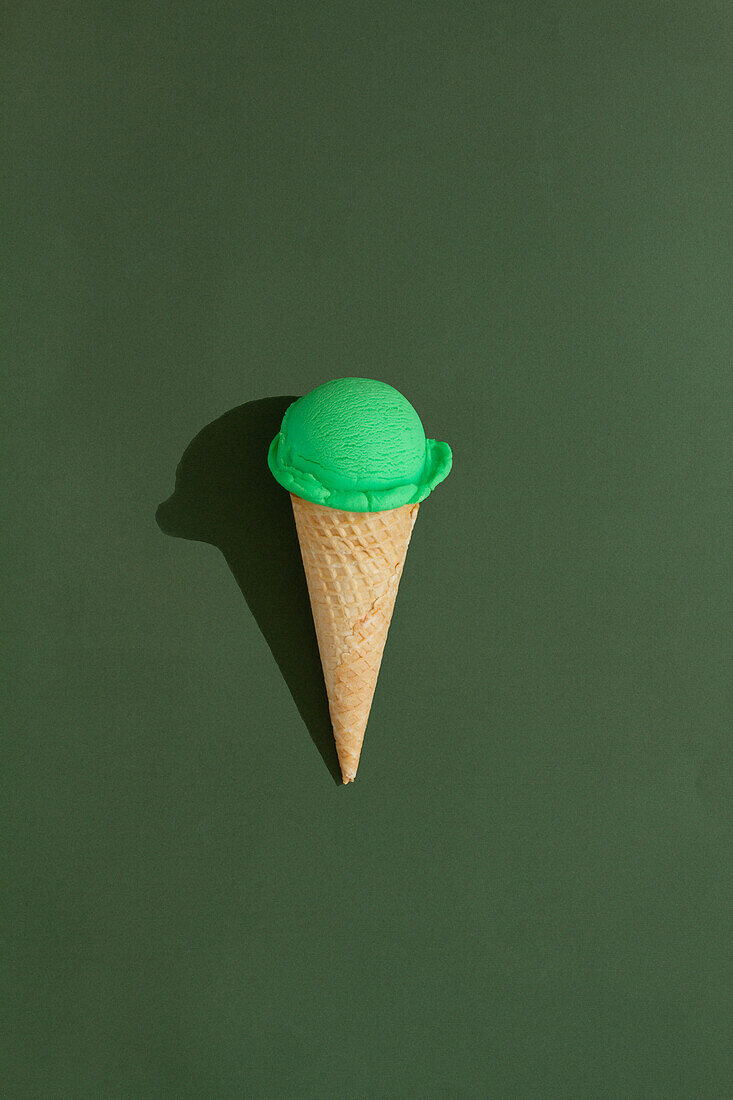 A vibrant green ice cream scoop sits atop a classic waffle cone against a green background, casting a playful shadow