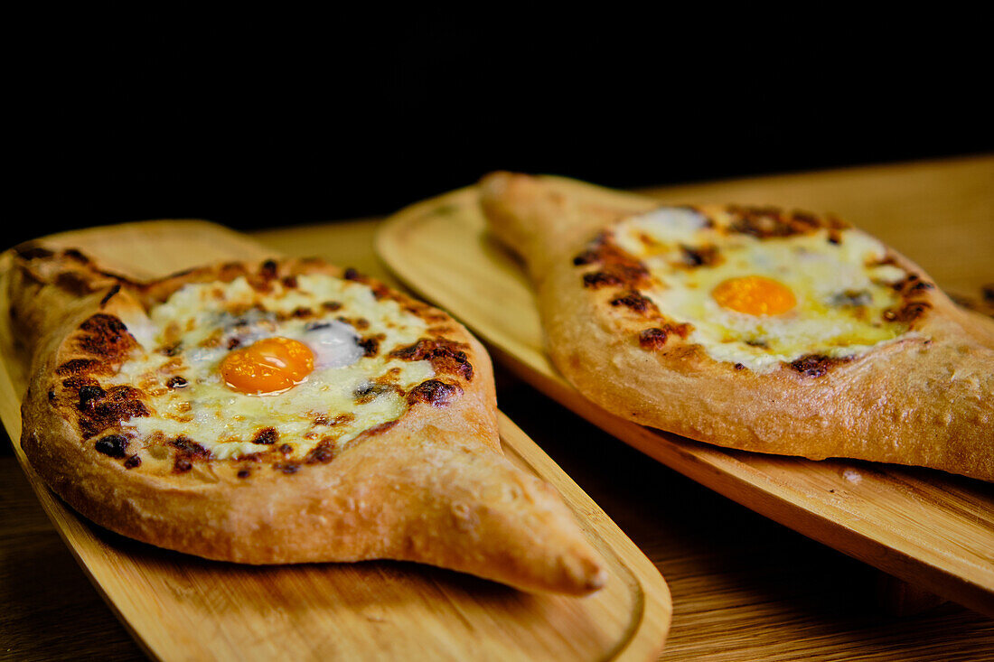 A closeup view of two traditional Georgian Khachapuris, each filled with melting cheese and a perfectly cooked egg, displayed aesthetically on rustic wooden boards