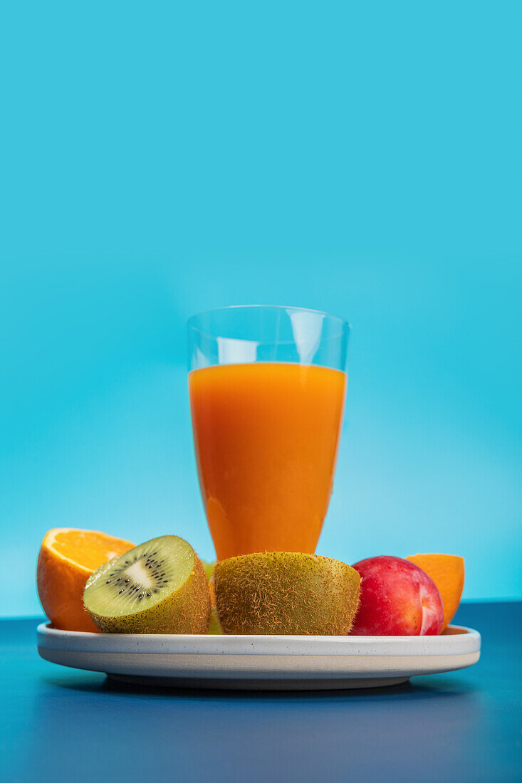 Ein lebhaftes Display mit Kiwi-, Orangen- und Pflaumenscheiben sowie einem frischen Glas Orangensaft vor einem markanten blauen Hintergrund. Ideal für Ernährungs- und Lifestyle-Themen.