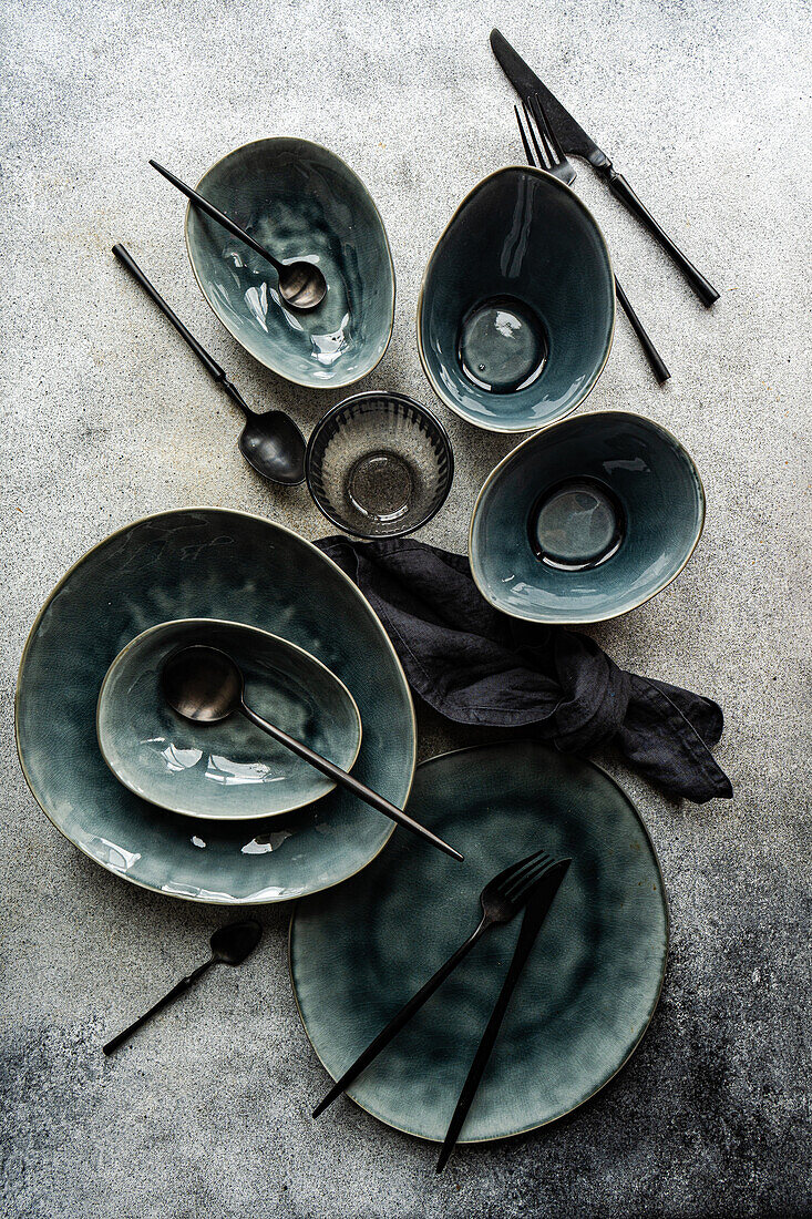 Top view of a luxurious table setting featuring emerald green ceramic plates, bowls, and dark cutlery on a textured grey surface.