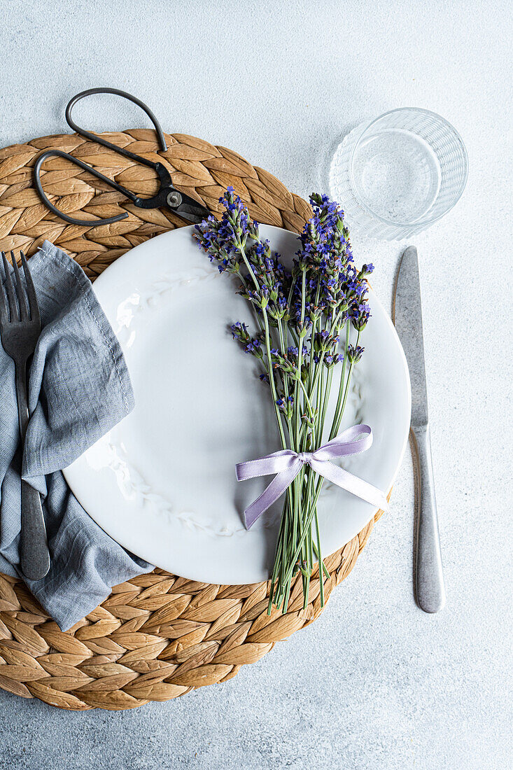 An elegant summer table setting featuring crisp white dishes, natural woven placemat, and fresh lavender tied with a ribbon, creates a serene dining atmosphere