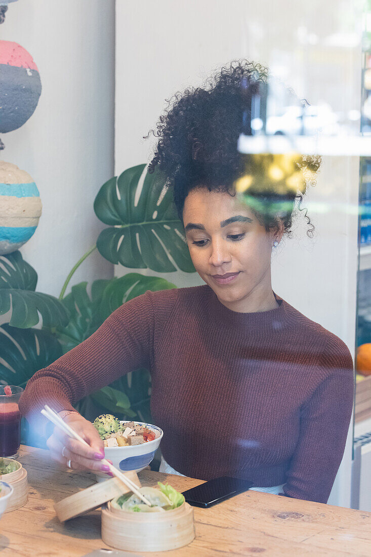 Zufriedene Afroamerikanerin sitzt am Tisch und isst ein appetitliches Poke-Gericht in einem Restaurant