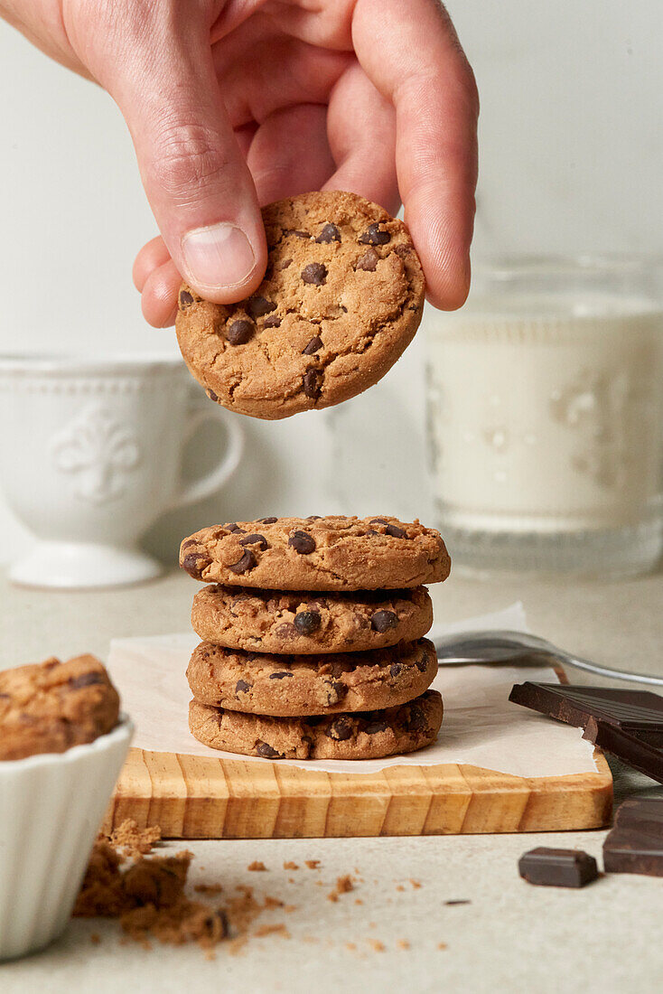 Anonymous person's hand grabbing a chocolate chip cookie from a delectable stack on a wooden cutting board, with milk and dark chocolate in the backdrop