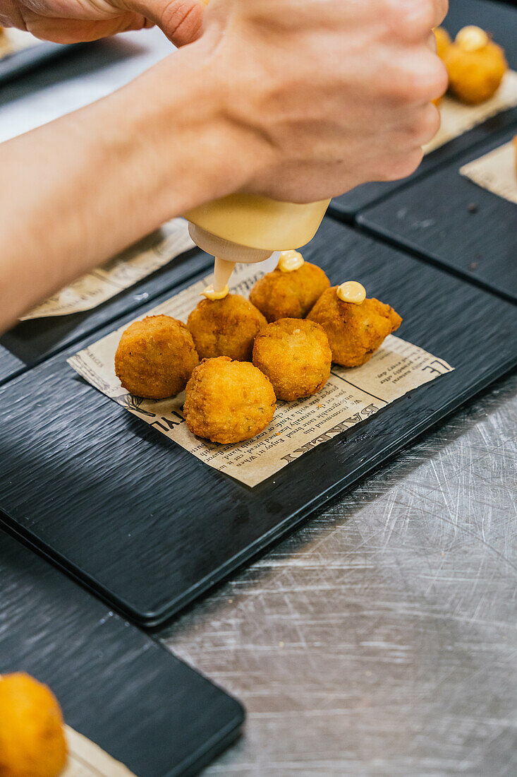 Anonymous chef hands meticulously drizzle a creamy sauce on golden croquettes arranged on a newspaper-lined serving tray