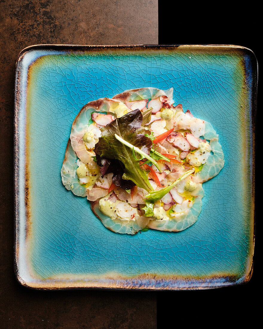 Ein appetitlicher Meeresfrüchtesalat mit gemischten Meeresfrüchten, frischem Salat und lebhaftem Gemüse, elegant serviert auf einem strukturierten blauen Keramikteller vor einem dunklen Hintergrund