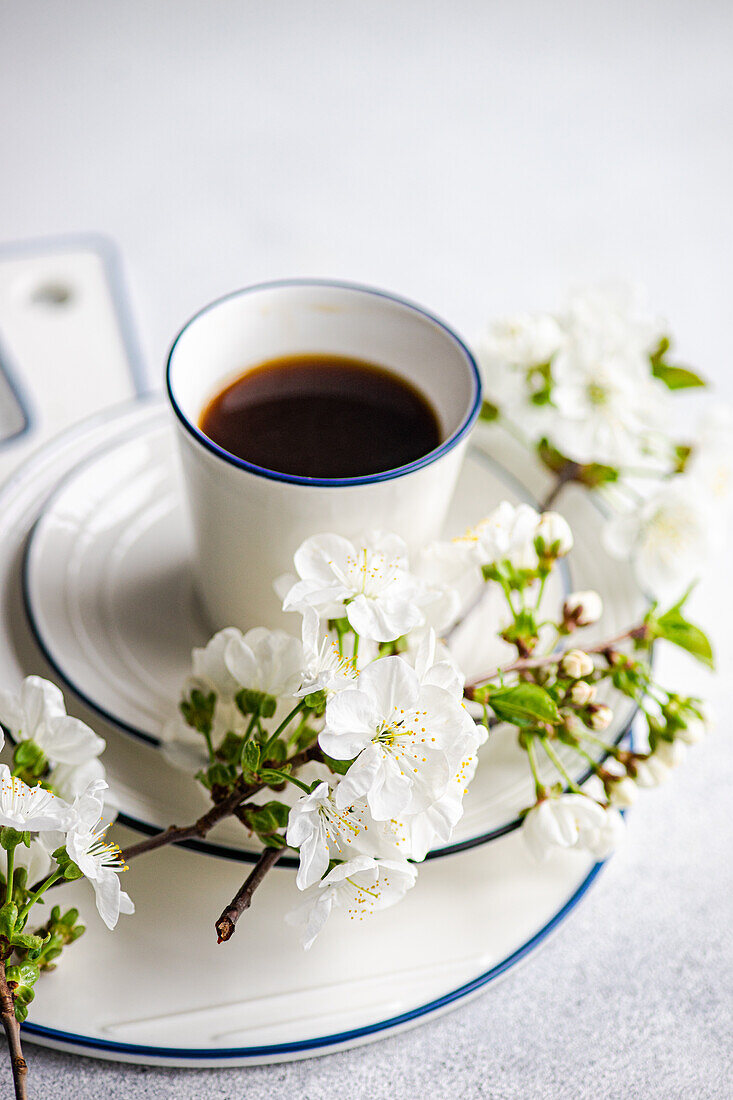 Ein weißer Emaillebecher, gefüllt mit schwarzem Kaffee, neben Kirschblüten auf einem hellen Hintergrund