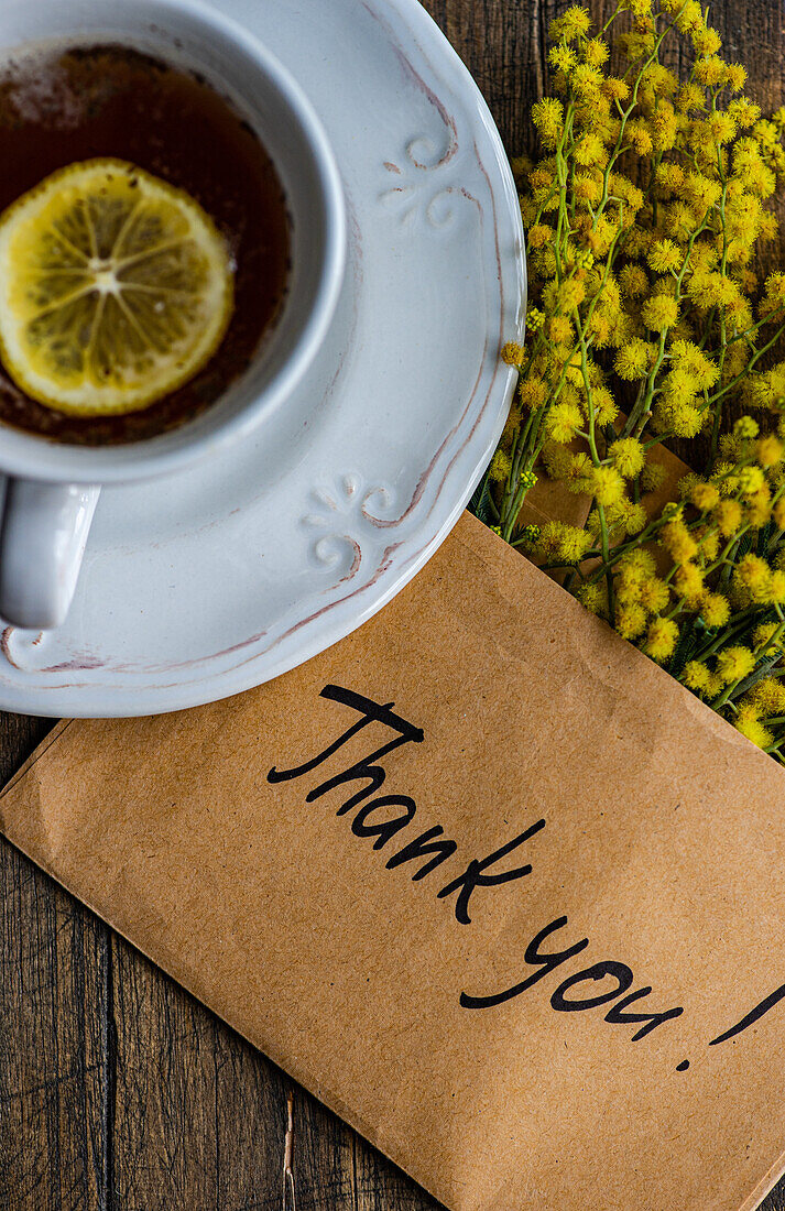 From above of a soothing tea with a lemon slice, paired with vibrant Mimosa flowers and a simple Thank You! note on rustic wood