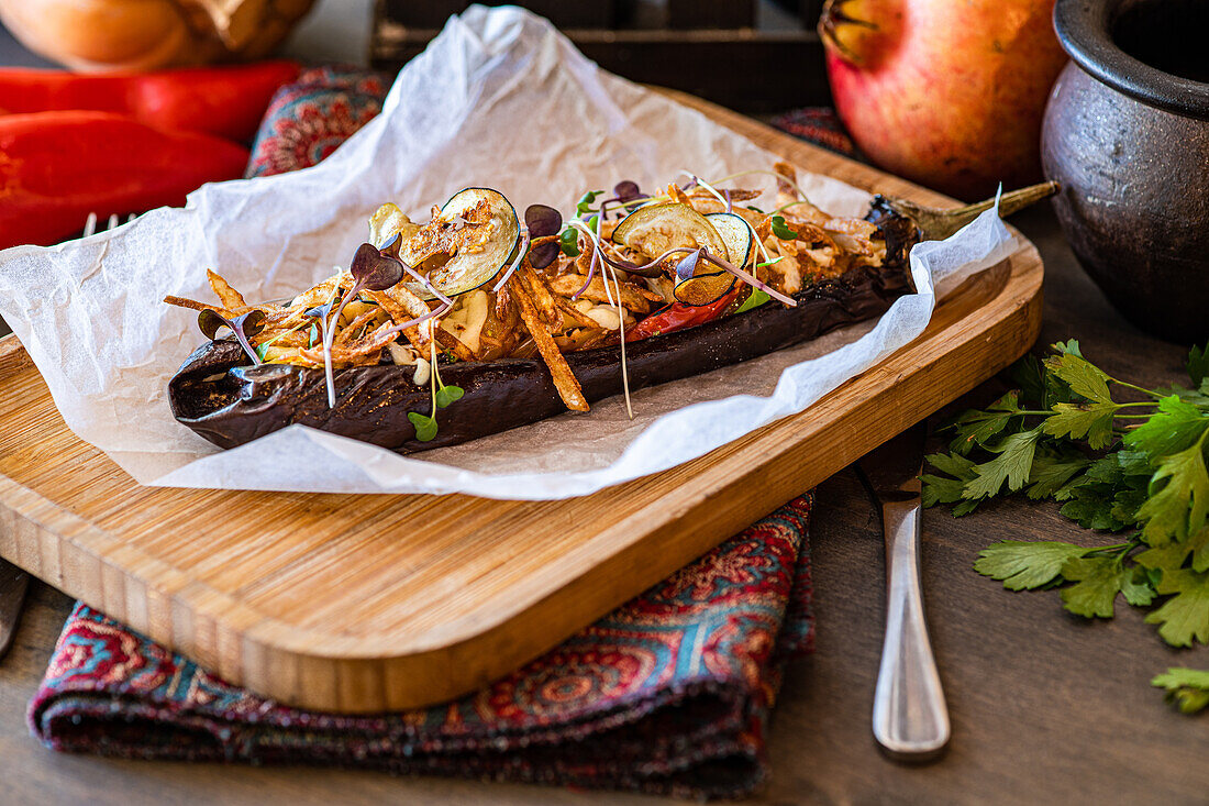 From above baked stuffed eggplant loaded with cherry tomatoes, bell pepper, onion, garlic, potato slices, and cheese, garnished with microgreens, served on a rustic wooden board with a decorative napkin.