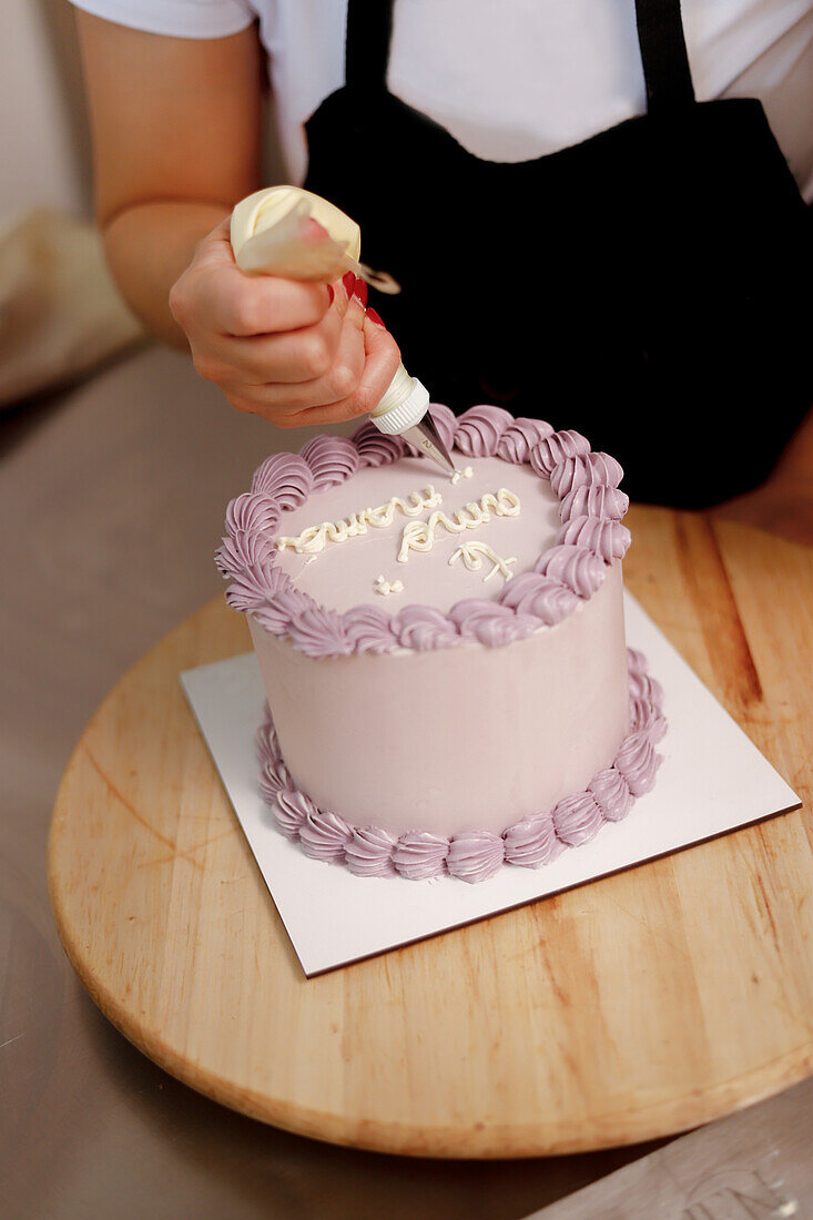 A baker decorates a cake with a pastry bag, adding elegant lettering to the smooth frosted surface