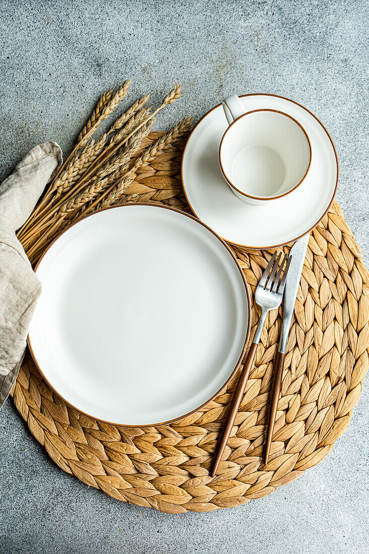 From above view of a stylish table setting featuring a bright white ceramic dinner plate, cup, and sleek silverware atop a woven placemat, accompanied by decorative wheat ears.