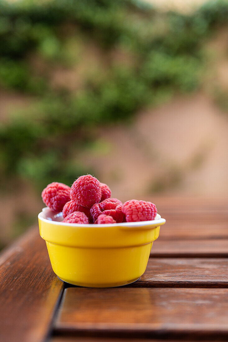 Vibrant red raspberries fill a cheerful yellow bowl, set on a wooden table, against a soft natural background