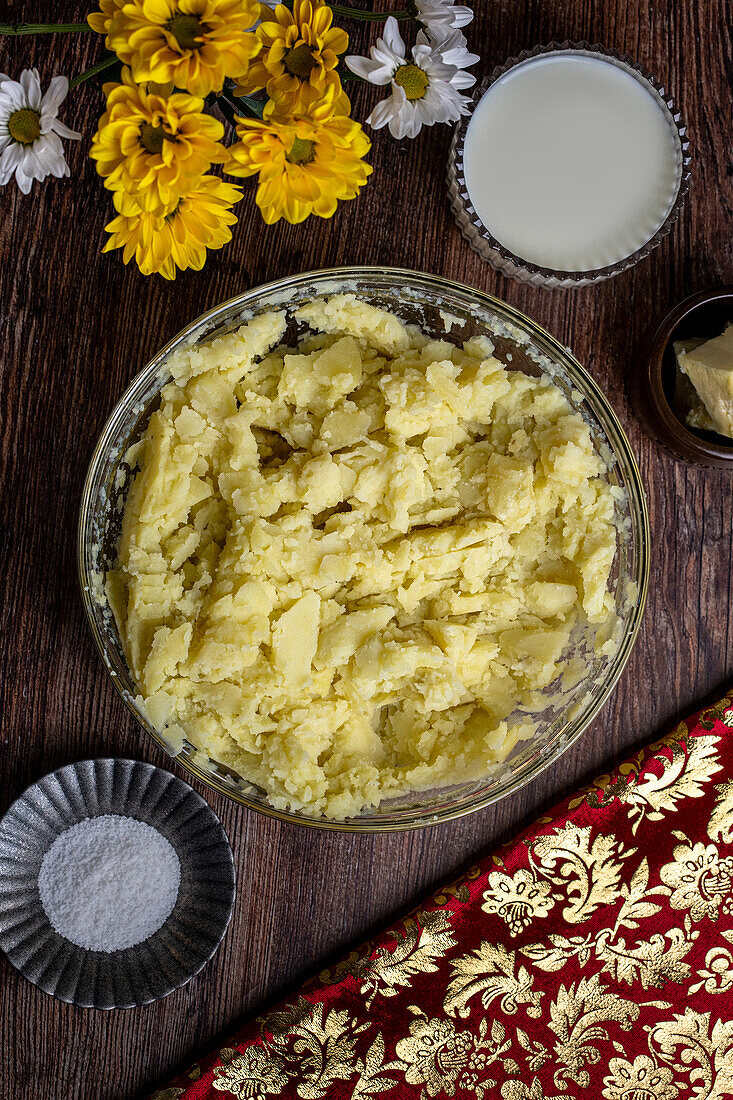 Draufsicht auf einen rustikalen Holztisch mit einer Schüssel mit hausgemachtem Kartoffelpüree, einer kleinen Schale mit Butter, einem Glas Milch und dekorativen Blumen, auf dem ein traditionelles hausgemachtes Mittagessen steht.