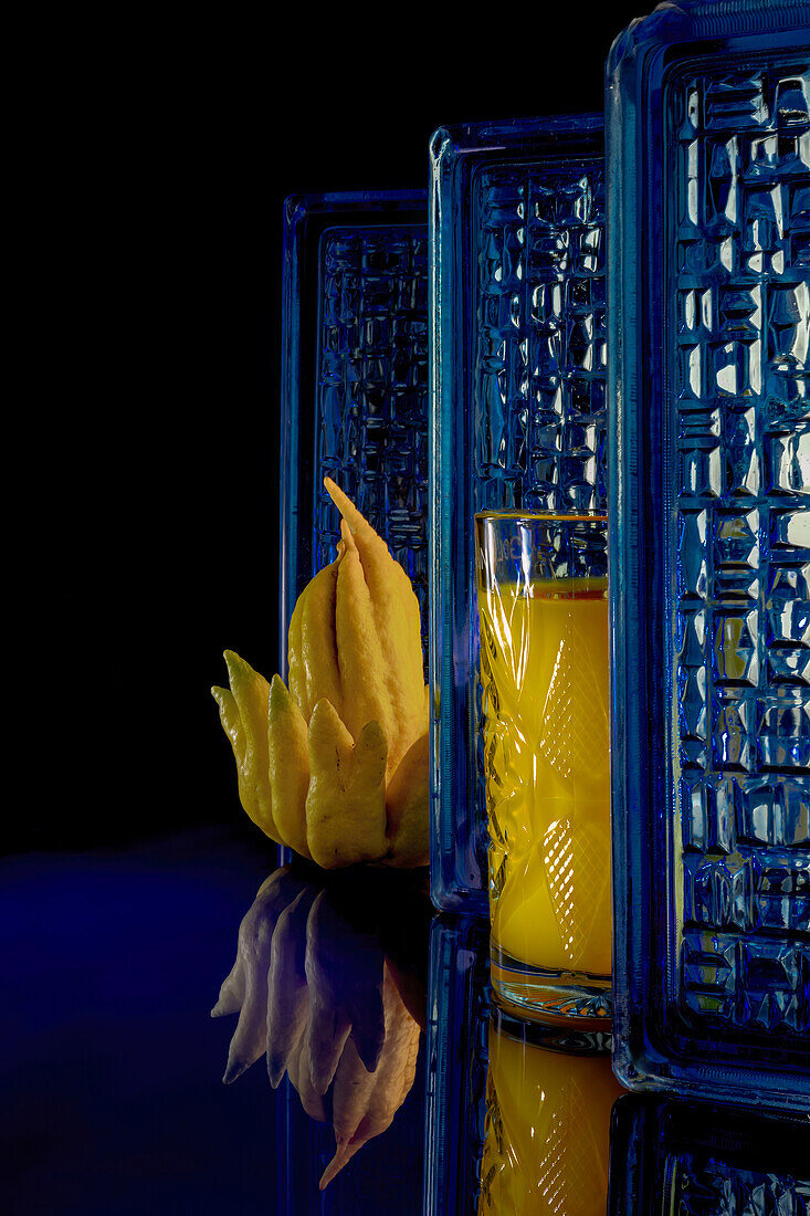 Ein lebhaftes Glas mit Mango- und Limettensaft neben einer einzigartigen Buddha-Handfrucht vor einem strukturierten blauen Hintergrund mit reflektierender Oberfläche