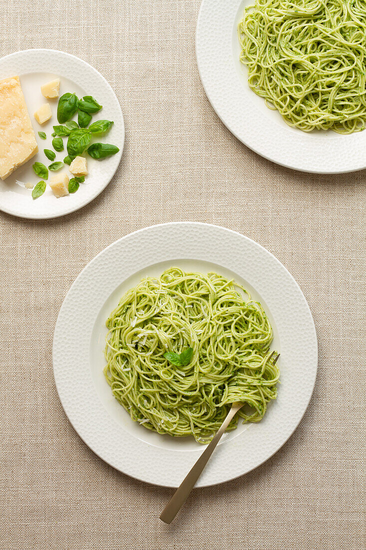 Draufsicht auf ein Spaghetti-Pesto-Gericht auf einem weißen Teller, begleitet von Parmesan und Basilikumblättern.