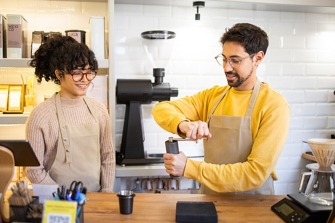 Two cheerful multiethnic baristas in aprons are happily engaging in their duties at a modern, well-lit coffee house, showcasing teamwork and customer service.