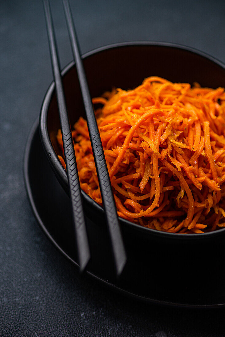 A close-up shot of a spicy Korean carrot salad, served in a black ceramic bowl with chopsticks on a dark background