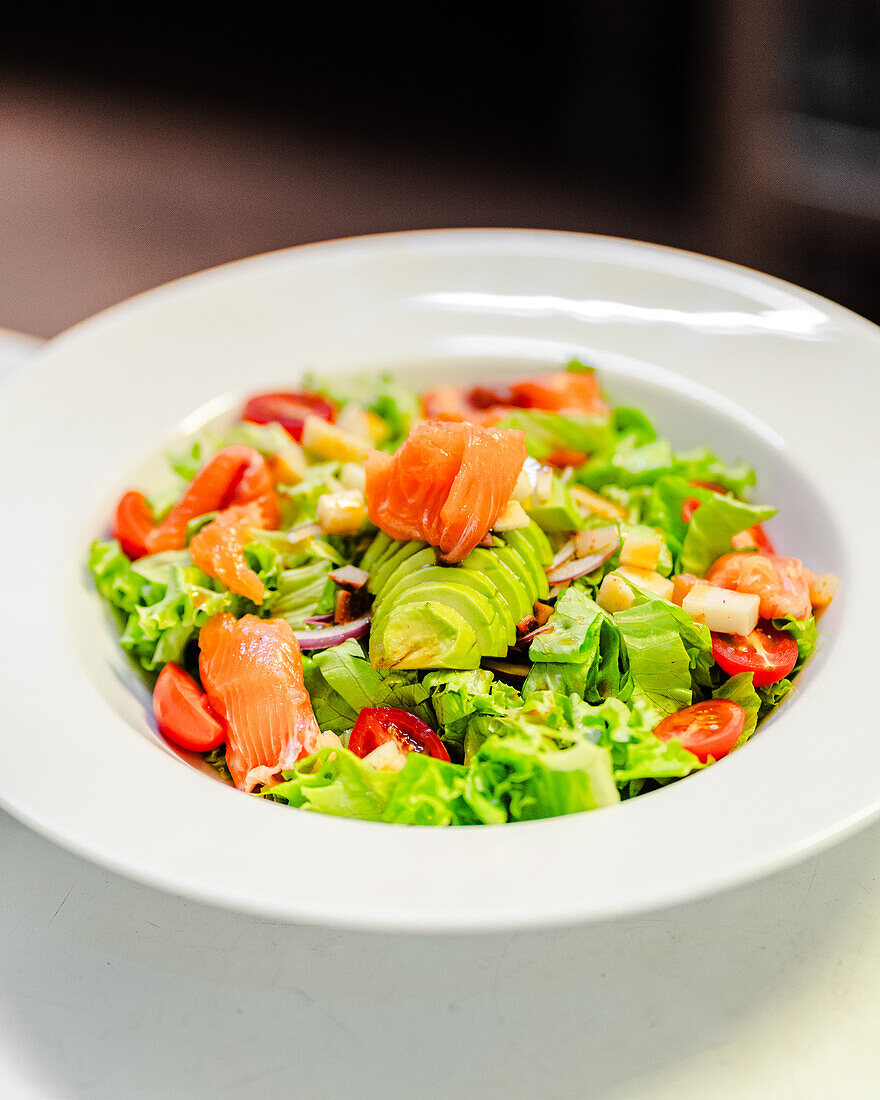 A vibrant house salad featuring fresh mezclum, succulent salmon slices, ripe cherry tomatoes, crisp onion, juicy apple, and creamy avocado served in a white bowl.