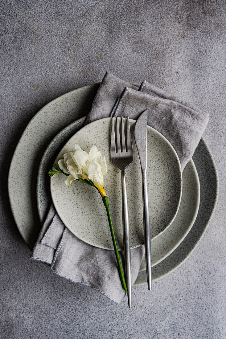 This image showcases a sophisticated table setting featuring a fresh freesia flower on speckled ceramic plates, complemented by sleek silverware and a gray linen napkin