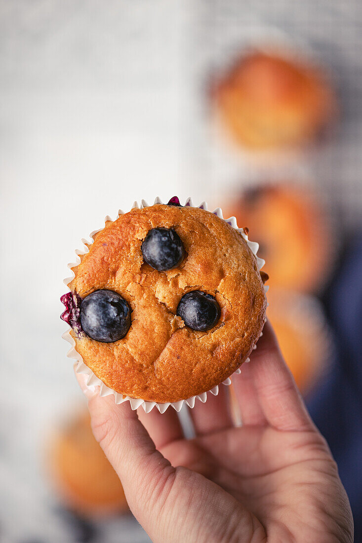 Nahaufnahme einer Hand, die einen leckeren Blaubeermuffin mit sichtbaren Beeren vor einem unscharfen Hintergrund hält