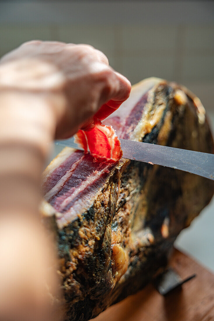 A close-up view of a hand slicing Serrano Ham off a leg mounted on a wooden stand with a sharp knife, highlighting the rich marbling and texture of the cured meat