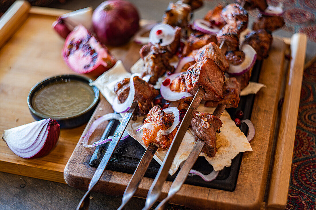 Eine rustikale Präsentation von georgischem Mzvadi mit saftigen gegrillten Fleischspießen, serviert auf einem Holzbrett mit geschnittenen Zwiebeln, Granatapfelkernen und traditionellem Fladenbrot - perfekt für kulinarische Themen