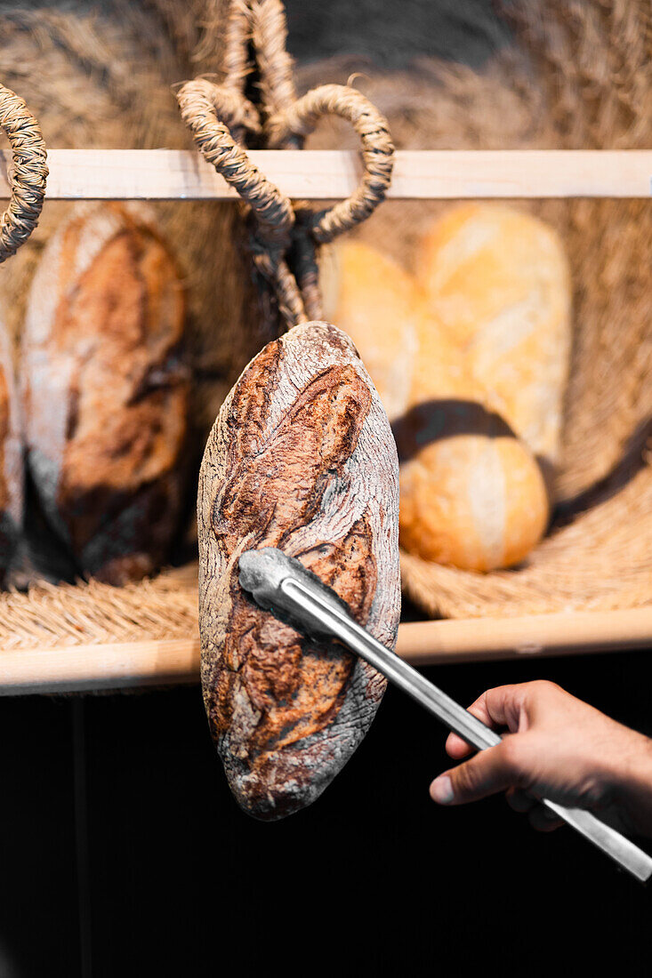 A cropped unrecognizable hand using tongs to pick a rustic sourdough bread loaf from a warm-hued selection displayed on a wooden shelf.