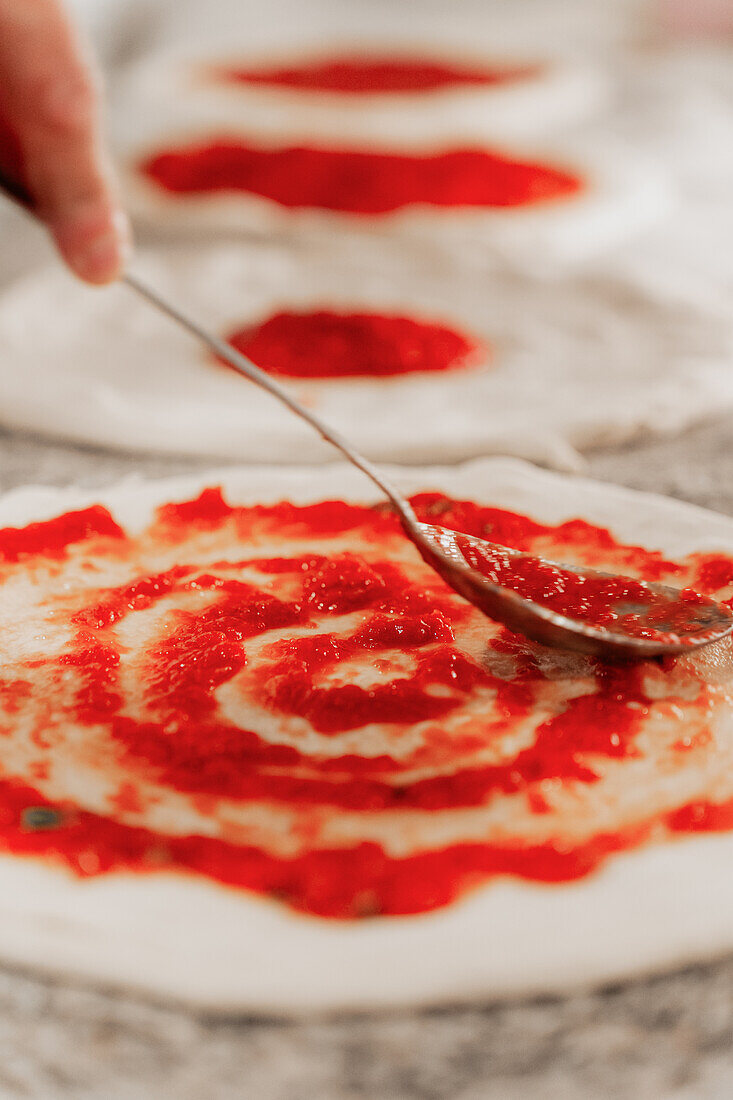 An anonymous chef in a pizzeria spreads tomato sauce on pizza dough with a ladle