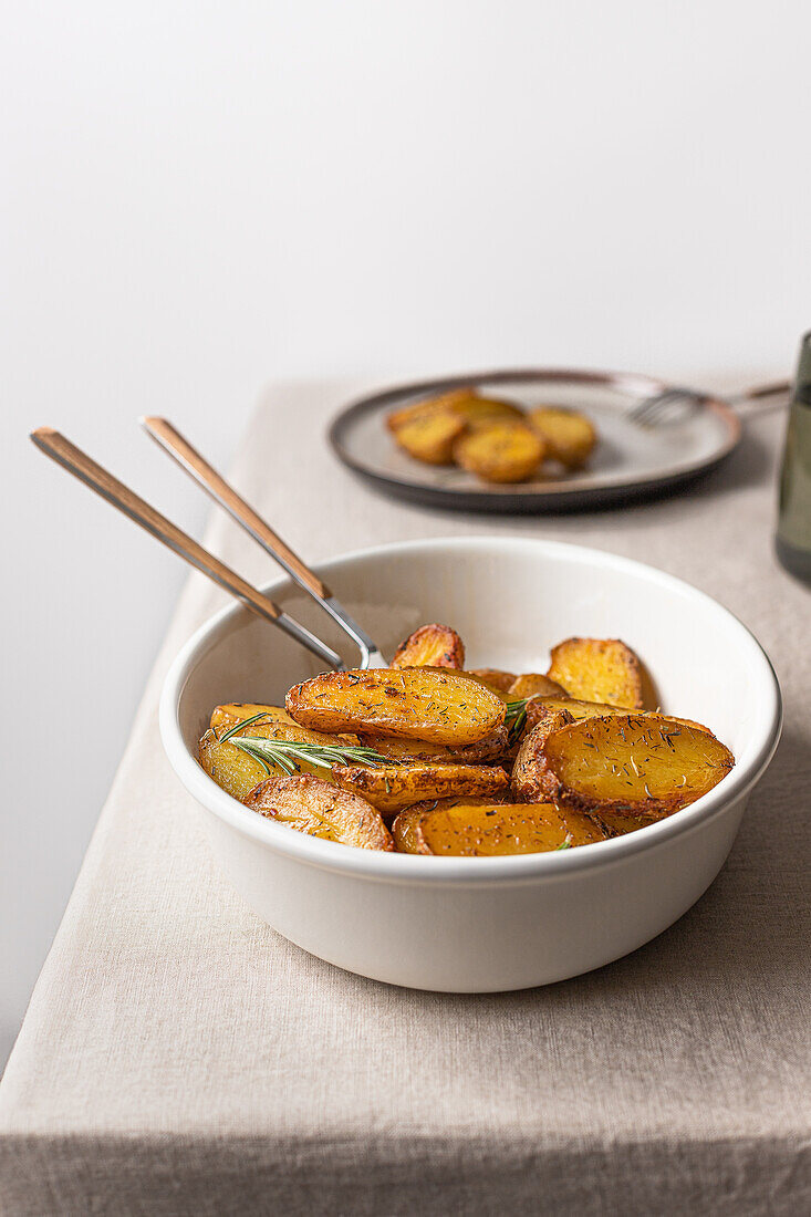 A white bowl filled with seasoned roasted potatoes, garnished with rosemary, on a linen-covered table with a secondary serving plate in the background.