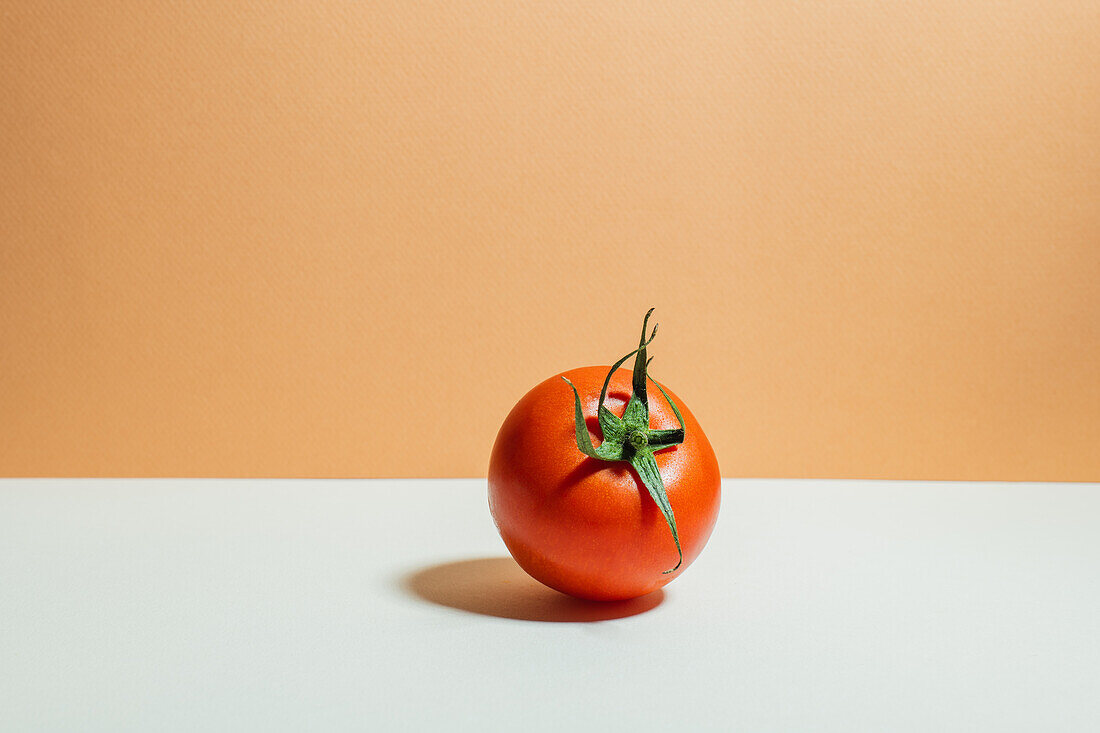 A ripe red tomato with a vibrant green stem placed against a white and orange two-tone background, showcasing simplicity and freshness.