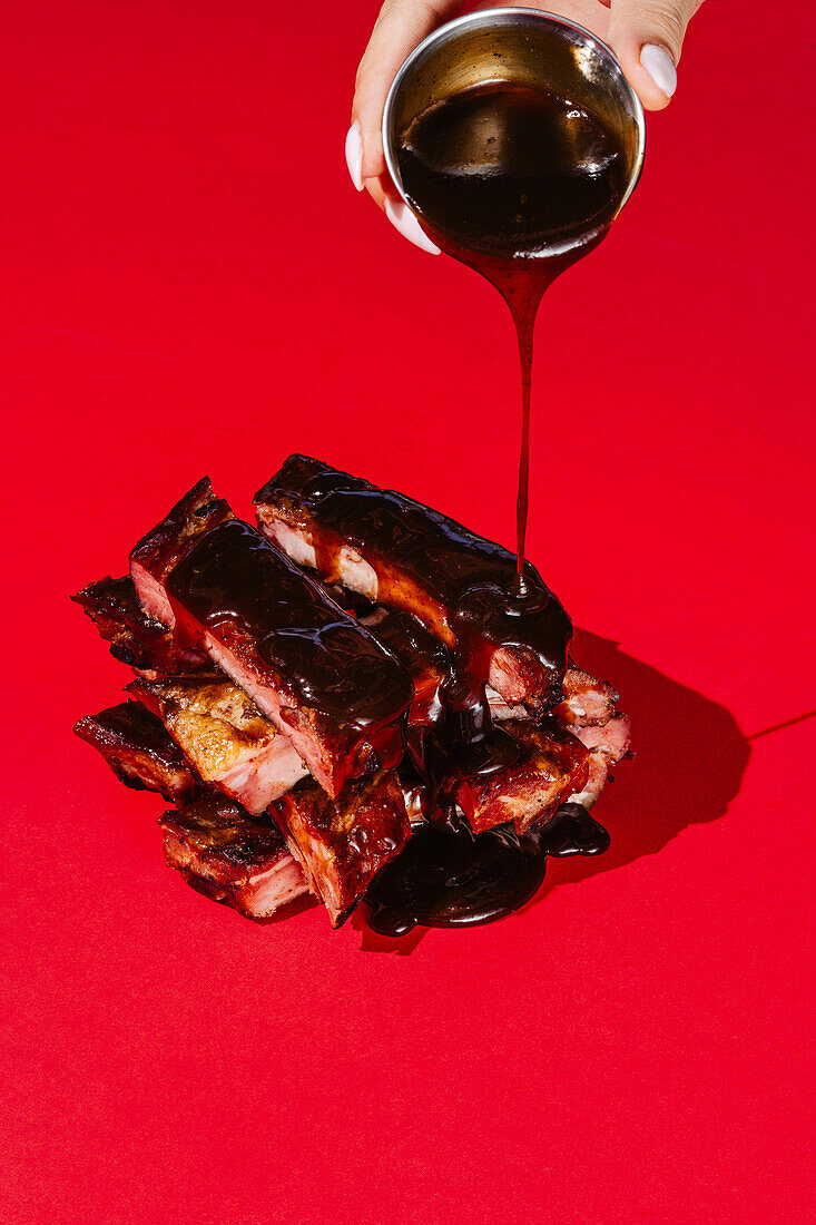 A succulent stack of grilled ribs is drenched in glossy barbecue sauce against a striking red background, highlighting a hand pouring the sauce from above