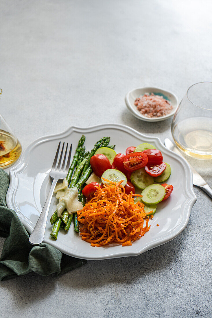 Top view of a nutritious vegetarian meal featuring grilled asparagus with melted cheese, sliced tomatoes and cucumbers, and grated carrots, lightly seasoned with oil and pink salt.