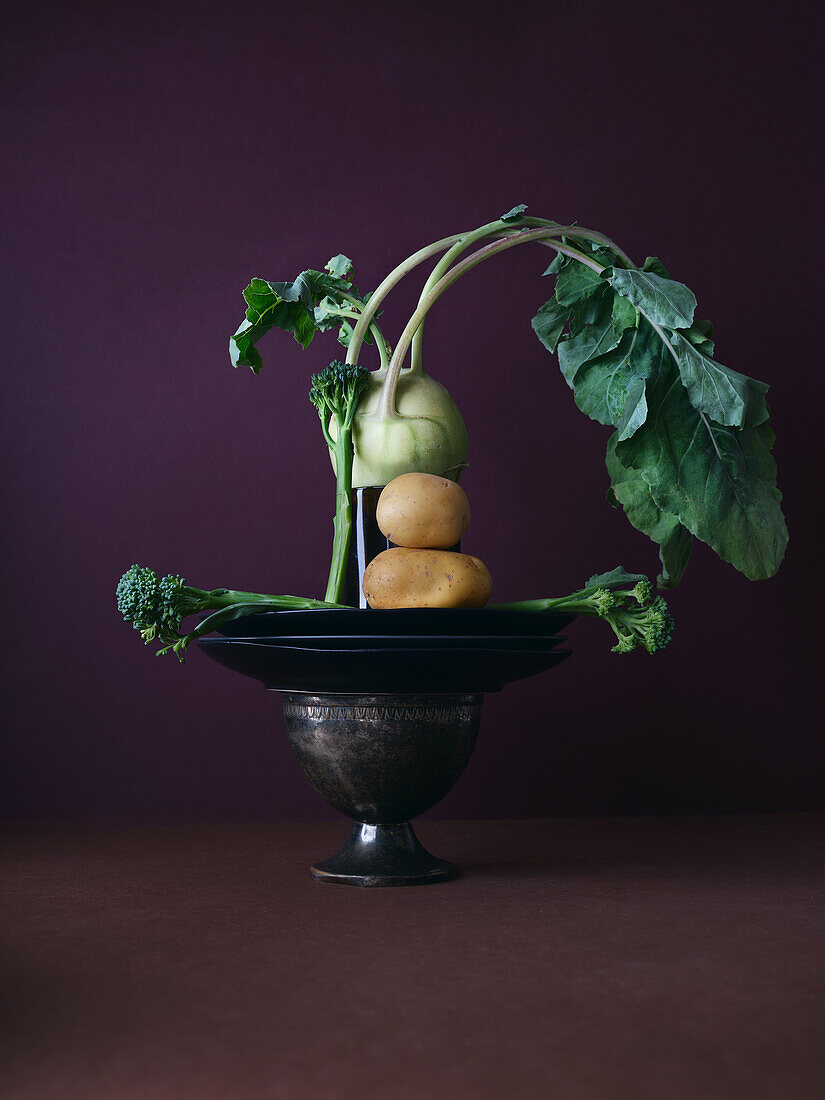 A creative arrangement of fresh vegetables balanced on an antique metal pedestal, featuring a kohlrabi, broccoli, and potatoes against a deep maroon backdrop