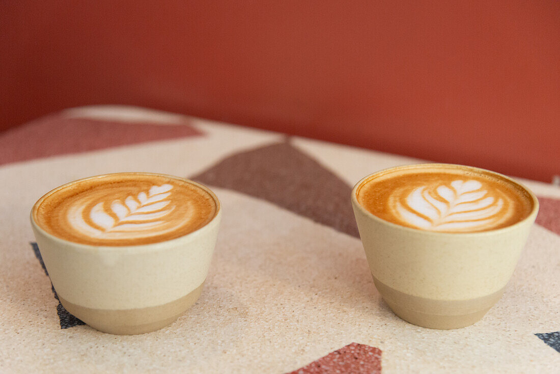 Dieses Bild zeigt zwei Tassen Kaffee mit kunstvoller Latte Art, die nebeneinander auf einem geometrisch gemusterten Tisch vor einem roten Hintergrund stehen.