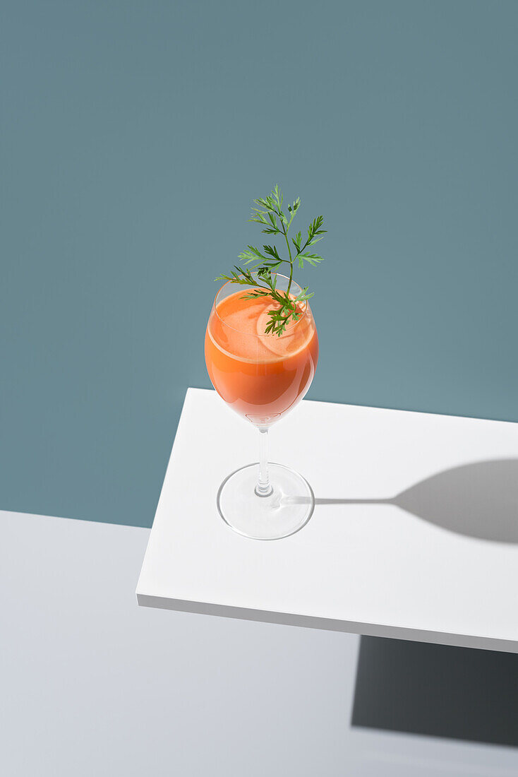 Minimalistische Präsentation eines Cocktails in einem Stielglas, verziert mit einem frischen grünen Zweig, vor einem modernen grauen Hintergrund mit einem starken Schatten.