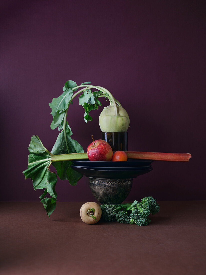 A creative display of healthy, fresh vegetables including broccoli, kohlrabi, apples, and a leafy stalk balancing on a metal bowl against a deep maroon background, suggesting composition and nutrition in May's peak season